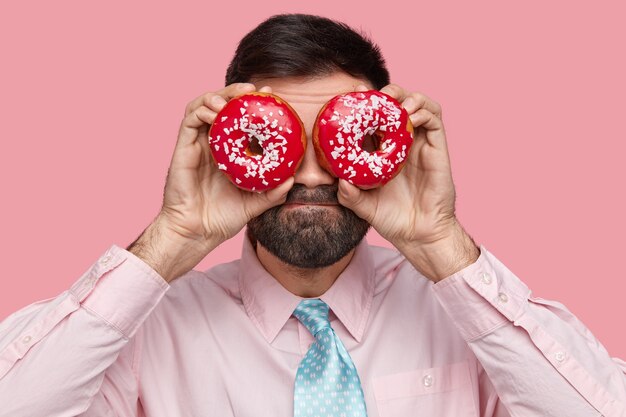 Attractive bearded man carries doughnuts near eyes, has dark stubble, dressed in formal outfit