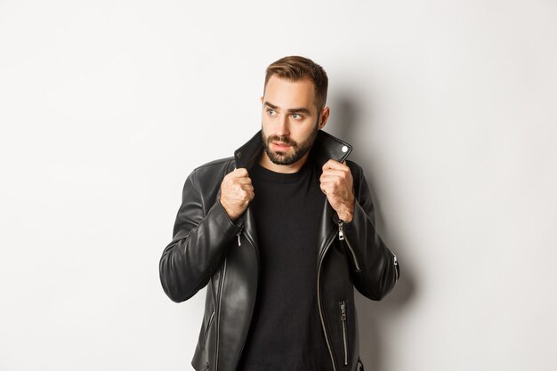 Attractive bearded man in leather biker jacket looking aside, standing confident