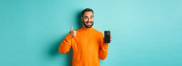 Attractive bearded man showing smartphone screen thumbs up recommending mobile app standing satisfie