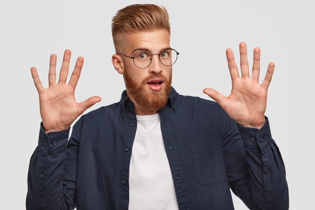 attractive bearded redhaired young male raises hands, shows palms, gestures actively, reacts on something terrible, dressed in casual clothes, poses against white wall.