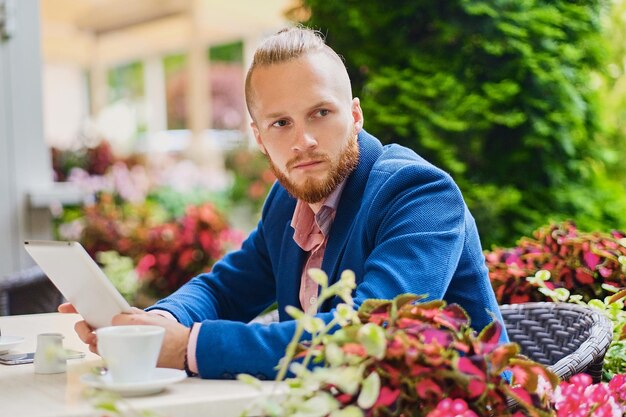 ピンクのシャツと青いジャケットを着た魅力的なあごひげを生やした赤毛の男性は、カフェのテーブルに座って、タブレットPCを使用しています。