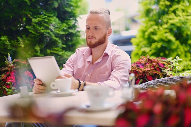 ピンクのシャツを着た魅力的なあごひげを生やした赤毛の男性は、カフェのテーブルに座って、タブレットPCを使用しています。