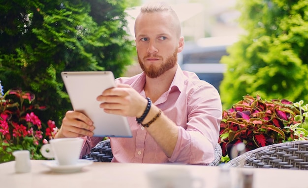 Attractive bearded redhead male in a pink shirt sits at the table in a cafe and using a tablet PC.
