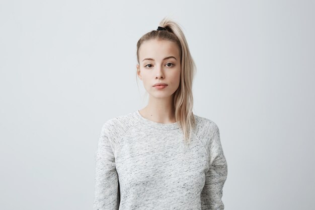 Attractive beautiful female with blonde pony tail, feeling self-assuarance while posing against blank wall.