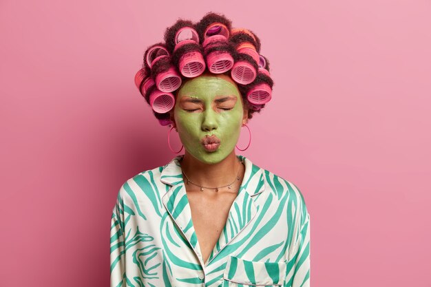 attractive beautiful young woman closes eyes, keeps lips rounded, applies green moisturizing mask on face, wears hair curlers, gets ready for special occasion, wears dressing gown