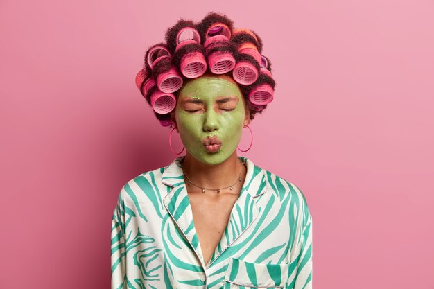 привлекательная красивая молодая женщина закрывает глаза, держит губы округлыми, наносит зеленую увлажняющую маску на лицо, носит бигуди, готовится к торжественному случаю, носит халат