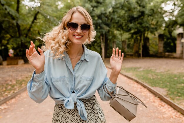 우아한 선글라스와 지갑을 입고 세련된 옷을 입고 공원에서 산책하는 매력적인 금발 솔직한 여자