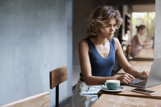 Attractive blond girl freelancer working laptop taking notes notebook sit alone cafe near window drink coffee, professional writer make post online blog, prepare files business meeting after lunch.