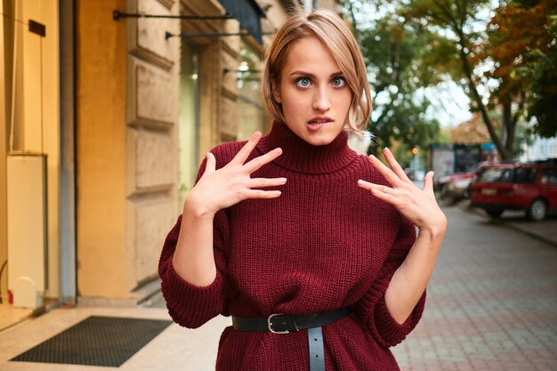 Attractive blond girl in knitted sweater playfully making faces fooling around on city street