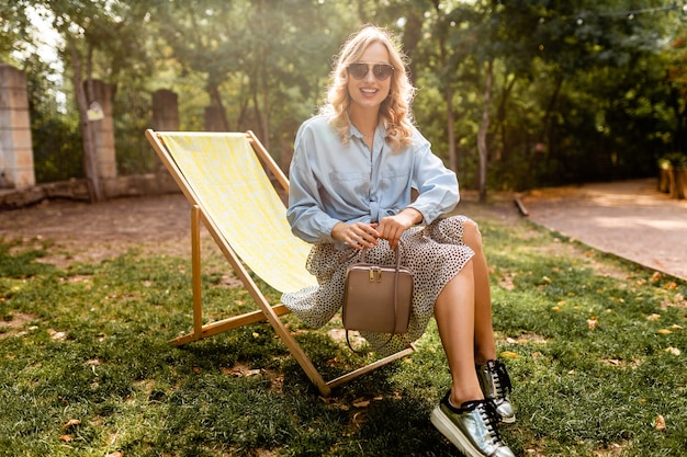 Attractive blond happy woman sitting relaxing in deck chair in summer outfit blue shirt, wearing silver sneakers, elegant sunglasses and purse