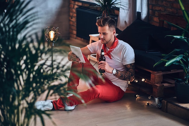 팔에 문신이 있는 매력적인 금발 남성, 로프트 인테리어가 있는 방에서 태블릿 PC를 사용하여 빨간 크리스마스 바지를 입고 있습니다.
