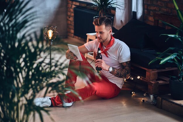 Attractive blond male with tattoos on arms, dressed in a red Christmas pants using tablet PC in a room with loft interior.