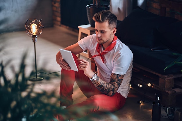 Attraente maschio biondo con tatuaggi sulle braccia, vestito con pantaloni rossi di natale utilizzando tablet pc in una stanza con interni soppalcati.