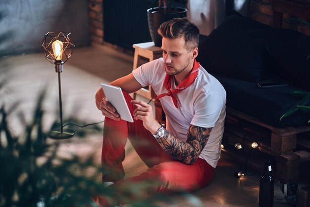 팔에 문신이 있는 매력적인 금발 남성, 로프트 인테리어가 있는 방에서 태블릿 PC를 사용하여 빨간 크리스마스 바지를 입고 있습니다.