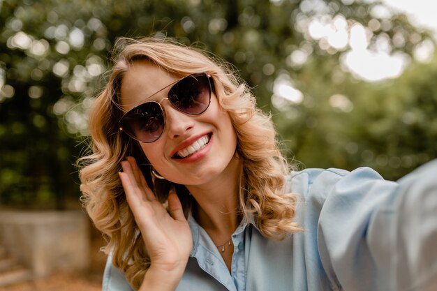 Attractive blond smiling white teeth woman walking in park in summer outfit taking selfie photo on phone