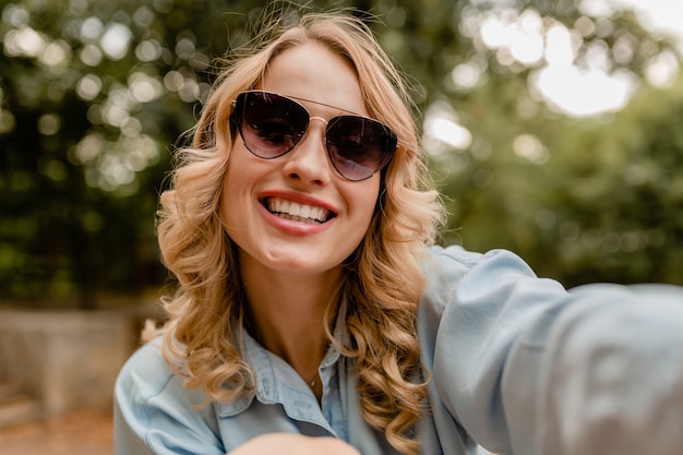 Attractive blond smiling white teeth woman walking in park in summer outfit taking selfie photo on phone