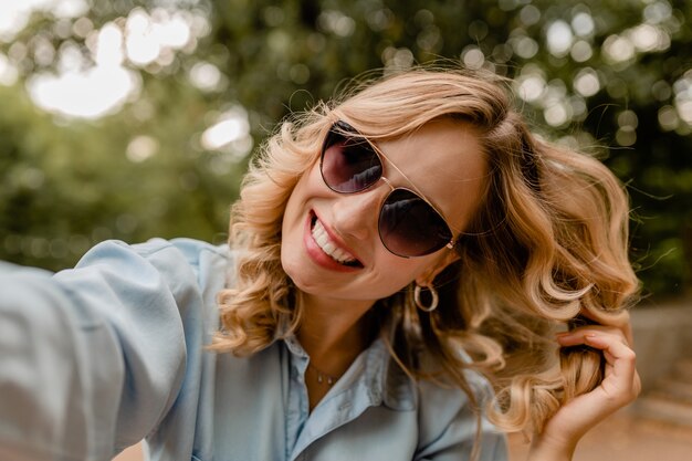 Attractive blond smiling white teeth woman walking in park in summer outfit taking selfie photo on phone