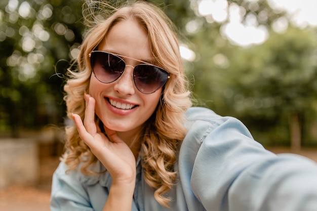 Attractive blond smiling white teeth woman walking in park in summer outfit taking selfie photo on phone