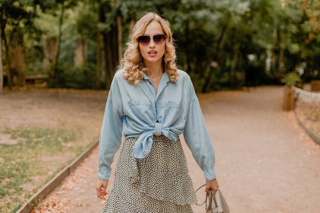 Attractive blond smiling woman walking in park in stylish outfit