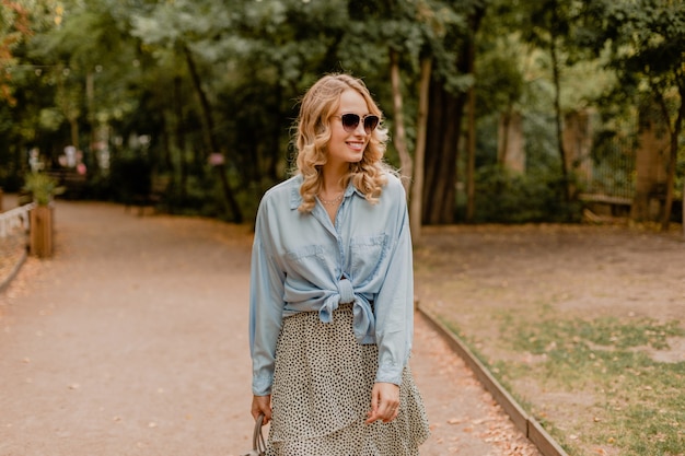 Attraente donna bionda sorridente che cammina nel parco in abito elegante