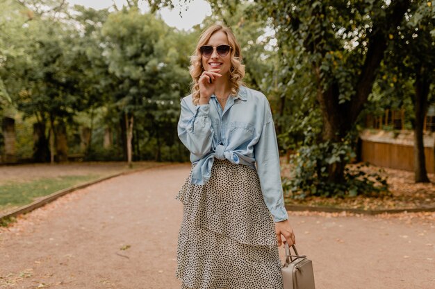Привлекательная белокурая улыбающаяся женщина гуляет в парке в стильной одежде
