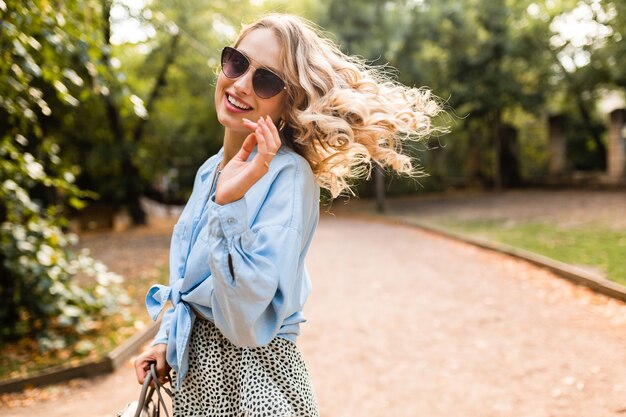Attractive blond smiling woman walking in park in summer outfit blue shirt and skirt, wearing elegant sunglasses and purse, street fashion style, happy mood