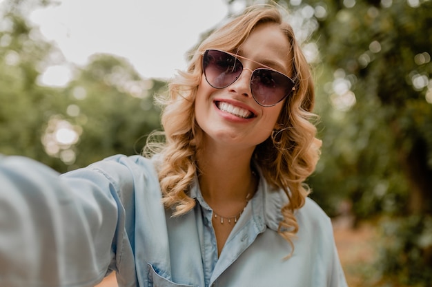 Attraente donna bionda sorridente che cammina nel parco in abito estivo prendendo foto selfie sul telefono