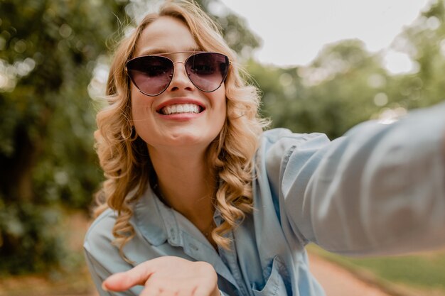 Attractive blond smiling woman walking in park in summer outfit taking selfie photo on phone