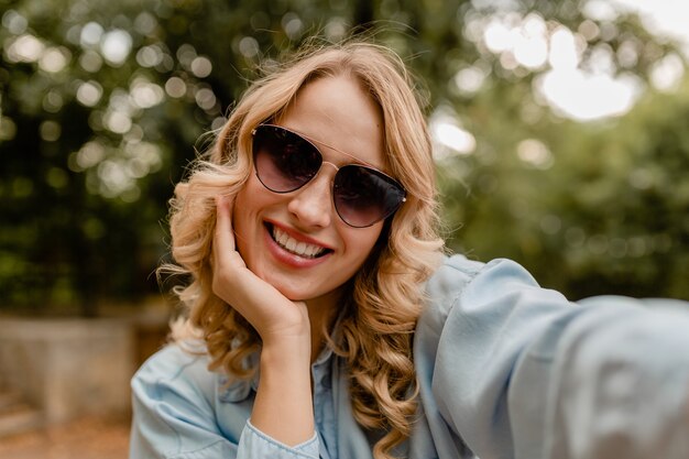 電話でselfie写真を撮る夏の衣装で公園を歩く魅力的な金髪の笑顔の女性