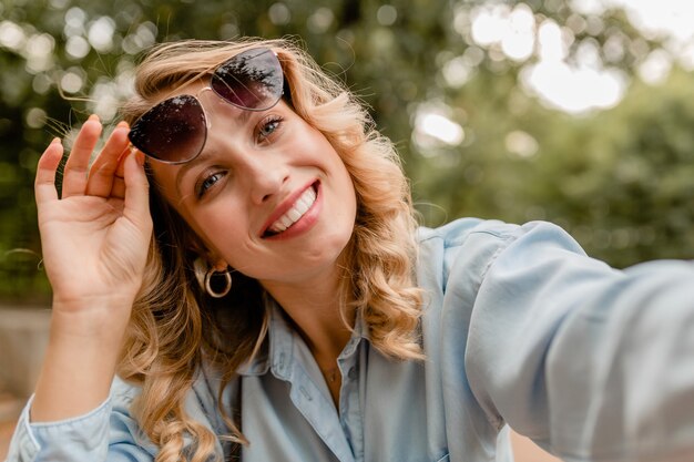 Attractive blond smiling woman walking in park in summer outfit taking selfie photo on phone