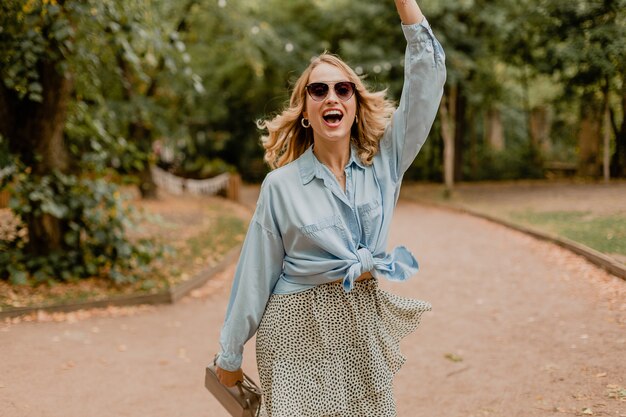 Attractive blond smiling woman waving hand hello walking in park in summer outfit