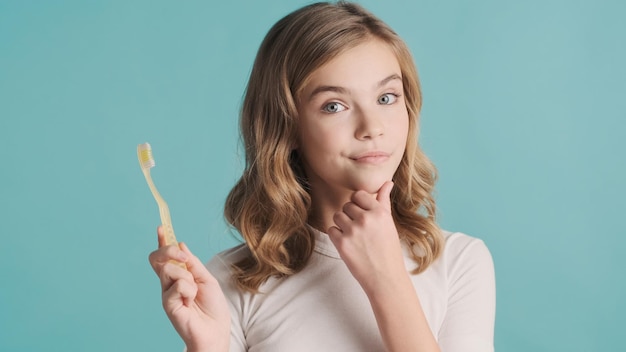 Attractive blond wavy haired teenager girl holding toothbrush looking thoughtful over blue background. Thinking expression