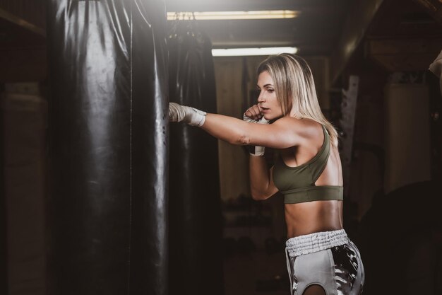 魅力的な金髪の女性は、キックボクシングスタジオでサンドバッグを使ったボクシングトレーニングを受けています。