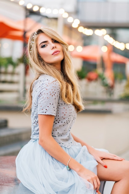 Attractive blonde girl in blue tulle skirt sitting on terrace background. She keeps hand on naked leg, looking to camera.