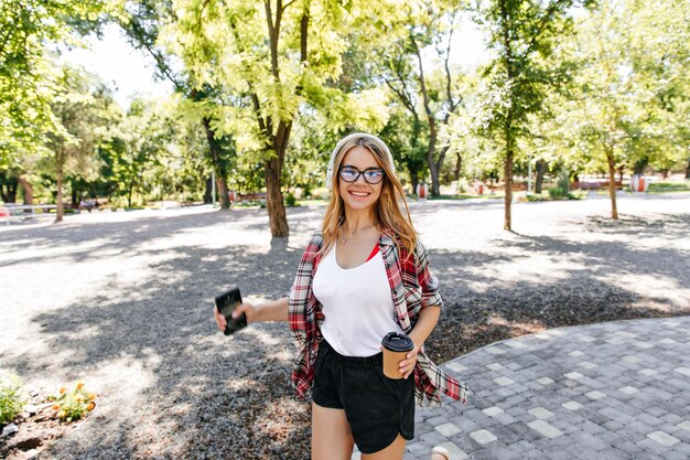 Привлекательная блондинка с чашкой кофе гуляет по парку Чудесная смеющаяся дама в наушниках позирует в солнечный день