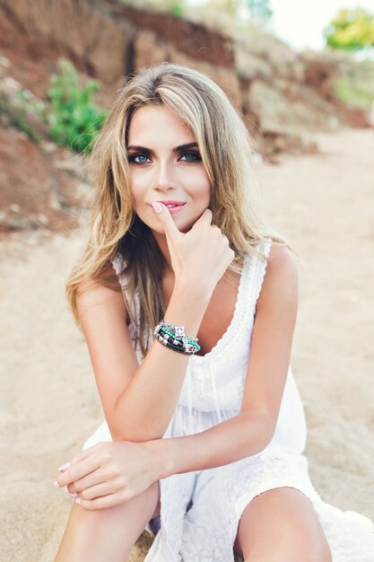 Attractive blonde girl with long hair and blue eyes is sitting on beach. She is looking to the camera.