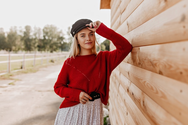 Attractive blonde with natural makeup looking with tenderness . Young woman in red pullover and stylish hat posing near the wooden house.