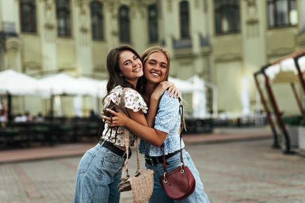 Attractive blonde woman in blue blouse and girl in white floral top hug outside Charming brunette lady in denim pants smiles and poses with friend outdoors