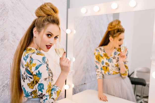 Attractive blonde young woman with luxury coiffure looking to camera with a glass of champagne behind the mirror in hairdresser salon