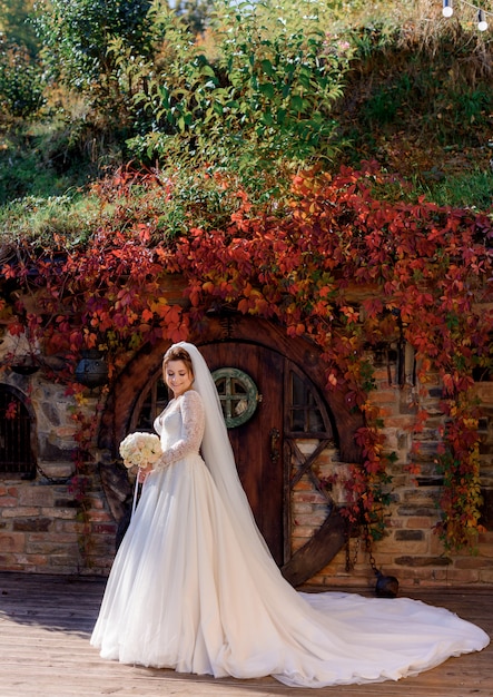 Attractive bride is standing in front of wooden entrance of a stone building with colorful leaves of ivy on the sunny day