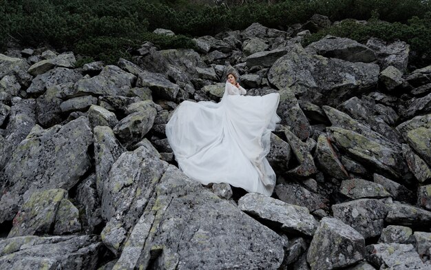 Attractive bride is standing on the huge grey rocks dressed in white ceremonial dress