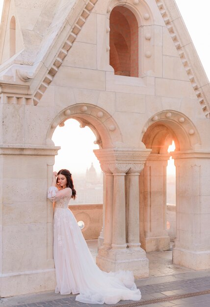 Attractive bride in  tender fashionable dress is standing near the stone column on a warm summer day