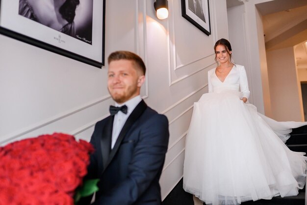 우아한 푹신한 웨딩 드레스를 입고 약혼자에게 계단을 내려가는 매력적인 신부 여자 여자 친구를 기다리는 빨간 장미 꽃의 큰 꽃다발을 가진 세련된 남자 결혼식 날 사랑하는 커플