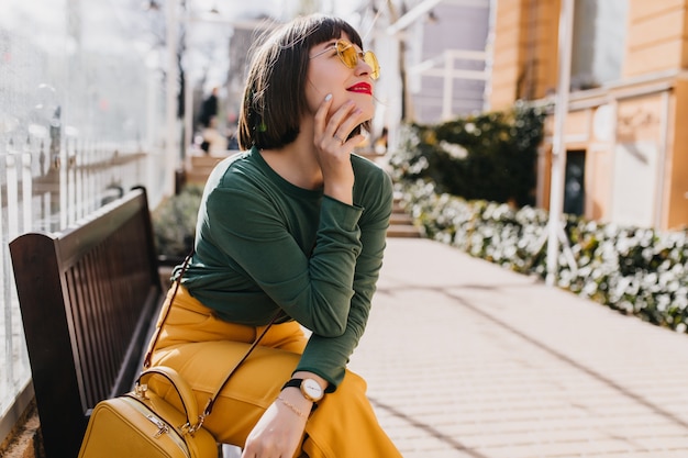 Attractive brown-haired girl in green sweater chilling on bench. Outdoor portrait of gorgeous lady in sunglasses dreamy posing on the street.