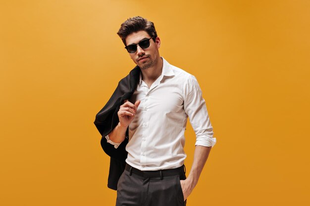 Attractive brunet man in stylish white shirt and trendy sunglasses poses on orange background and holds black jacket