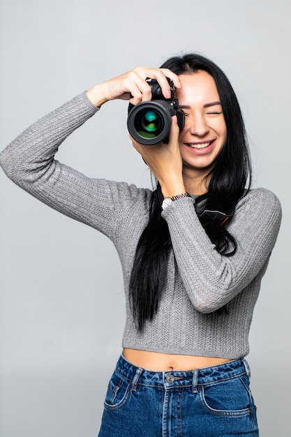 Attractive brunette aims her camera. composing a photograph in studio, isolated on gray wall