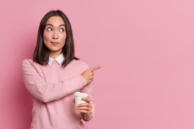 Attractive brunette Asian woman holds disposable cup of coffee points aside and shows blank space advertises place for promotion dressed in casual clothes suggests visit cafe and grab some drink