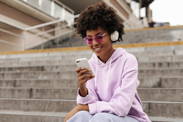 Attractive brunette curly woman in purple hoodie and pink sunglasses smiles, holds phone, listens to music in headphones and sits on stairs outdoors