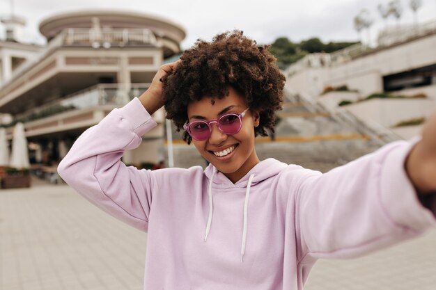 Attractive brunette curly woman in purple hoodie, pink sunglasses smiles sincerely, rejoices and takes selfie outside