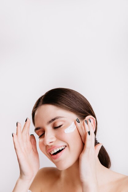 Attractive brunette enjoying spa procedure on isolated wall. Smiling girl applies moisturizer.